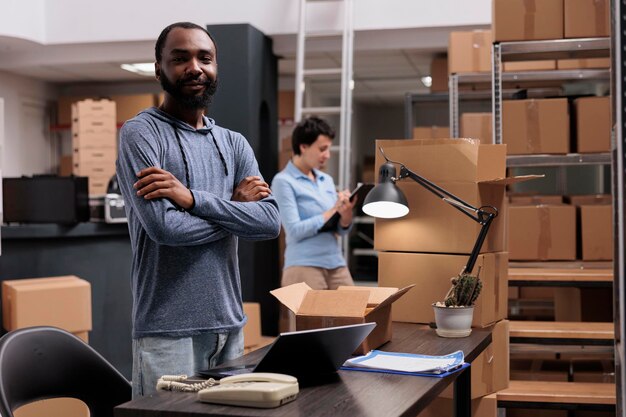 Ritratto di dipendente afroamericano in piedi con il braccio incrociato in magazzino mentre lavora alla consegna dei pacchi ai clienti, preparando scatole di cartone. Società di adempimento del centro di distribuzione