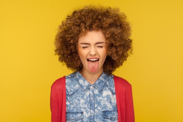 Ritratto di disobbediente pazza donna divertente con i capelli ricci in abito casual che mostra la lingua fuori e ha chiuso gli occhi dispiaciuto comportandosi indisciplinato cattivo studio indoor girato isolato su sfondo giallo