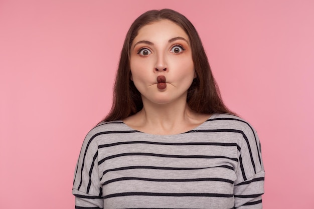 Ritratto di divertente donna meravigliata in felpa a righe che fa una smorfia di faccia di pesce con le labbra e sembra stupita confusa con un'espressione sciocca idiota. studio indoor girato isolato su sfondo rosa