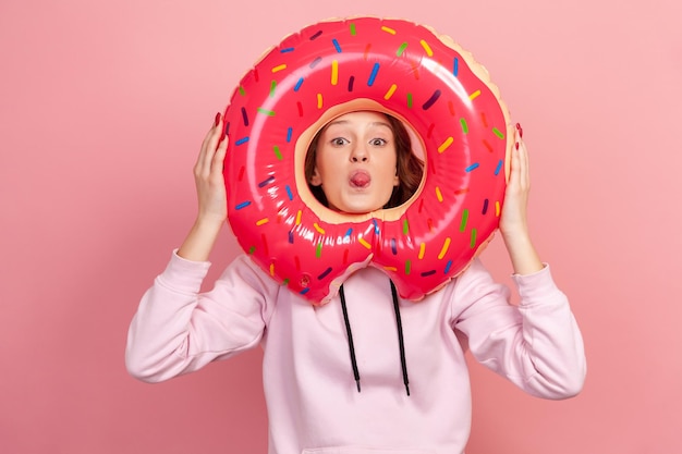 Ritratto di divertente ragazza adolescente dai capelli ricci in felpa con cappuccio che mostra la lingua attraverso l'anello di gomma rosa della ciambella che tiene nelle mani, scherzando, riposa. Studio indoor girato isolato su sfondo rosa