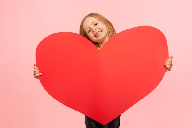 Ritratto di dolce bambina allegra che fa capolino dal grande simbolo del cuore rosso e sorride alla telecamera congratulandosi per la festa della mamma dicendo ti amo girato in studio al coperto isolato su sfondo rosa