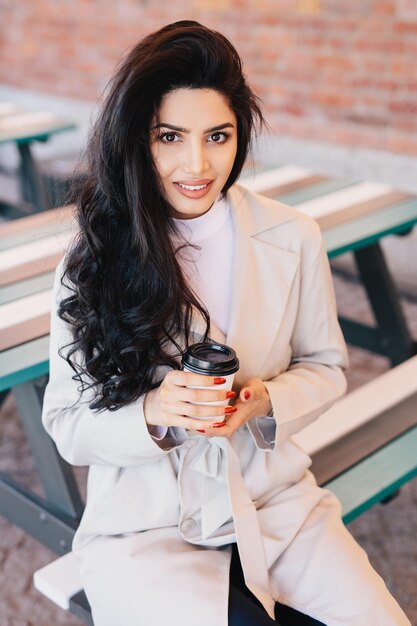 Ritratto di donna adorabile con lunghi capelli ondulati scuri occhi luminosi e labbra carnose che indossa un camice bianco seduto alla panchina in un caffè all'aperto che beve caffè da asporto in posa a porte chiuse sorridendo piacevolmente