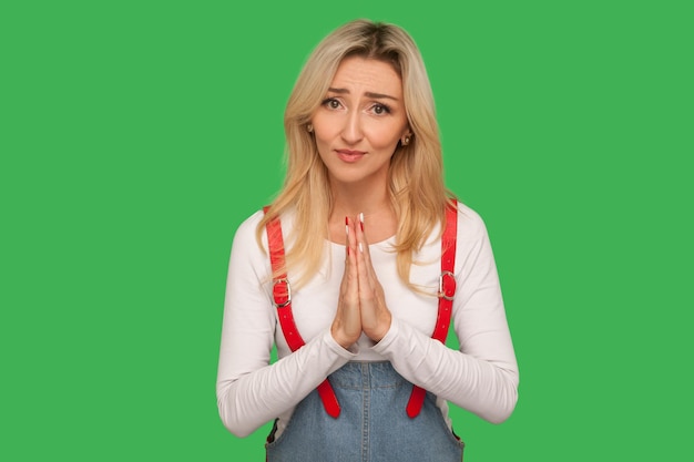 Ritratto di donna adulta implorante preoccupata in tuta di jeans che prega per aiuto facendo appello con l'espressione colpevole di scusarsi sinceramente dicendo per favore indoor studio shot isolato su sfondo verde