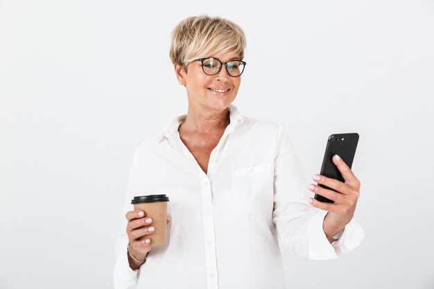 Ritratto di donna adulta sorridente che indossa occhiali tenendo il cellulare e la tazza di caffè da asporto isolata sul muro bianco in studio