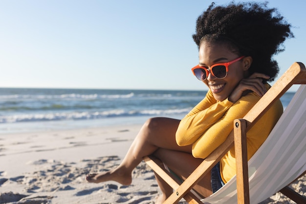 Ritratto di donna afro-americana in occhiali da sole seduta sulla sedia a sdraio sorridente sulla spiaggia soleggiata in riva al mare. Estate, relax, tempo libero e vacanza, inalterati.