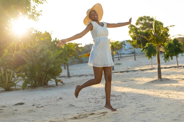 Ritratto di donna afroamericana felice in cappello da sole e abito bianco da spiaggia che balla sulla spiaggia soleggiata. Estate, libertà, relax e vacanze, inalterate.