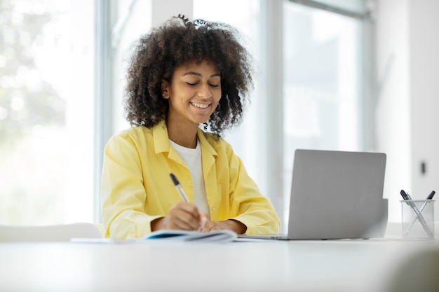 Ritratto di donna afroamericana seduta al tavolo, utilizzando il laptop e scrivendo sul taccuino, studiando