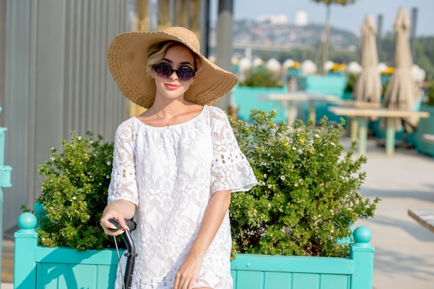 Ritratto di donna alla moda, bella ragazza bionda in abito bianco, occhiali da sole e cappello a cavallo scooter