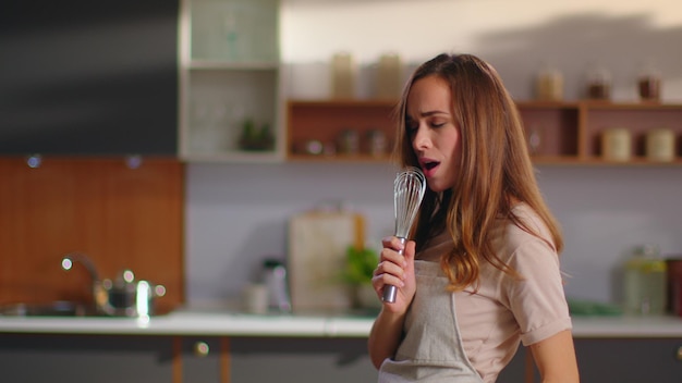 Ritratto di donna allegra che canta una canzone nei baffi sulla cucina domestica al rallentatore Ragazza positiva che si diverte con un utensile da cucina a casa Giovane donna che balla sulla cucina