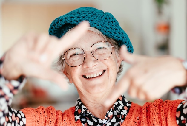 Ritratto di donna anziana attraente felice sorridente che indossa berretto e occhiali che gesturing il telaio della fotocamera con le mani che guardano la fotocamera