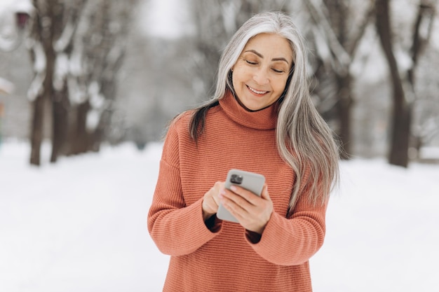 Ritratto di donna anziana con capelli grigi in maglione lavorato a maglia che parla al telefono su sfondo invernale