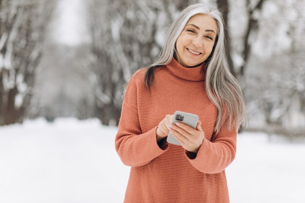 Ritratto di donna anziana con capelli grigi in maglione lavorato a maglia che parla al telefono su sfondo invernale