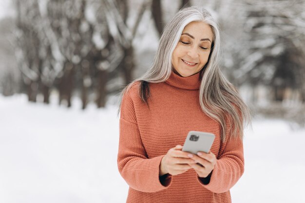 Ritratto di donna anziana con capelli grigi in maglione lavorato a maglia che parla al telefono su sfondo invernale