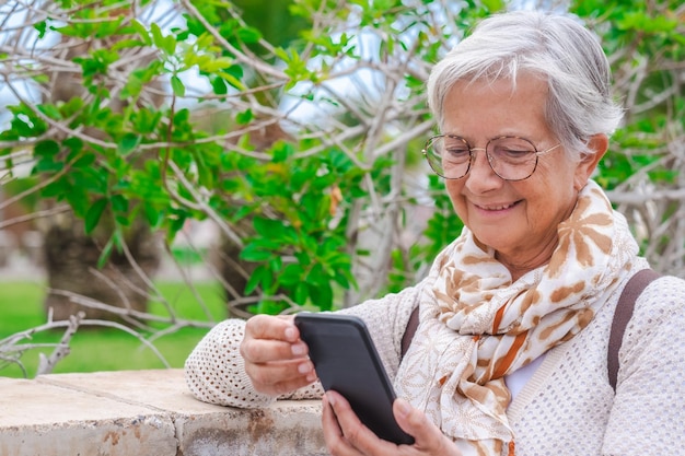 Ritratto di donna anziana dai capelli bianchi sorridente che utilizza il telefono cellulare bianco in piedi sul parco pubblico godendo della pensione o delle vacanze Signora anziana che parla tramite smartphone Sfondo di foglie verdi