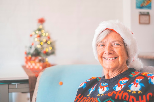 Ritratto di donna anziana in pensione sorridente con cappello da Babbo Natale e vestiti caldi che celebrano il natale a casa. Vecchia santa femmina sorridente mentre guarda la fotocamera. Signora anziana che si gode le vacanze di Natale