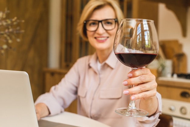 Ritratto di donna anziana matura sorridente che tiene in mano un bicchiere di vino mentre si utilizza il computer portatile sul tavolo della cucina. Freelance che lavora a casa concetto.