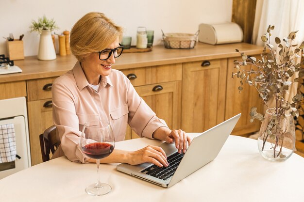 Ritratto di donna anziana matura sorridente che tiene in mano un bicchiere di vino mentre si utilizza il computer portatile sul tavolo della cucina. Freelance che lavora a casa concetto.