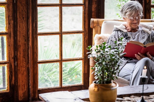 Ritratto di donna anziana seduta a casa sulla poltrona leggendo un libro che indossa un maglione caldo e occhiali da vista. Soggiorno confortevole, finestra in legno