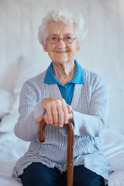 Ritratto di donna anziana seduta e sorriso sul letto con il bastone da passeggio della felicità e relax nella casa di cura