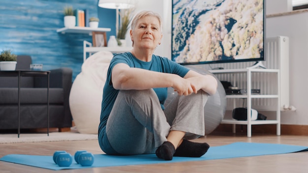 Ritratto di donna anziana seduta sul tappetino da yoga a casa. Persona anziana che guarda la telecamera, pronta a fare esercizio fisico e attività con attrezzature per l'allenamento. Formazione pensionati