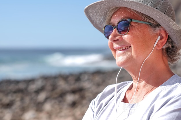 Ritratto di donna anziana sorridente attraente che indossa cappello e occhiali da sole mentre ascolta musica seduta sulla spiaggia Orizzonte sull'acqua
