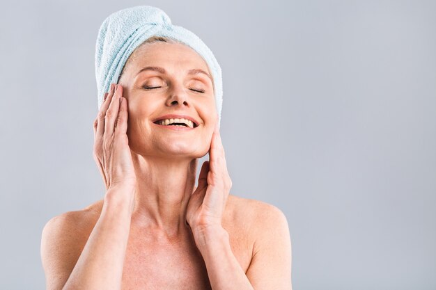 ritratto di donna anziana sorridente che sente la pelle morbida con la mano dopo il bagno termale donna matura avvolta in un asciugamano sul corpo e capelli bagnati guardando la fotocamera