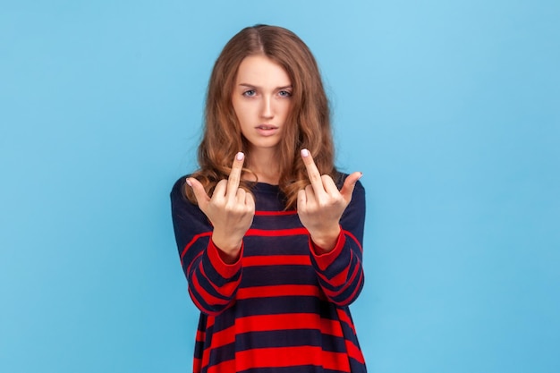 Ritratto di donna arrabbiata pazza che indossa un maglione in stile casual a righe che mostra il dito medio gesto maleducato viso pieno di odio e risentimento Studio interno girato isolato su sfondo blu