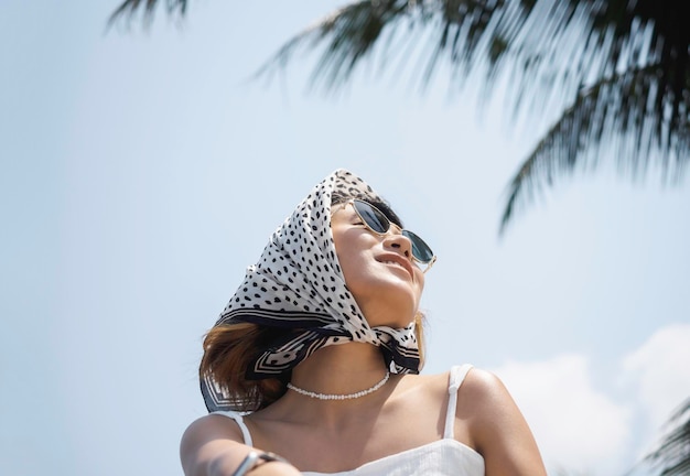 Ritratto di donna asiatica abbastanza felice in camicia bianca casual che indossa occhiali da sole e sciarpa per capelli goditi il sole in spiaggia sotto le palme da cocco e lo sfondo del cielo blu in estate