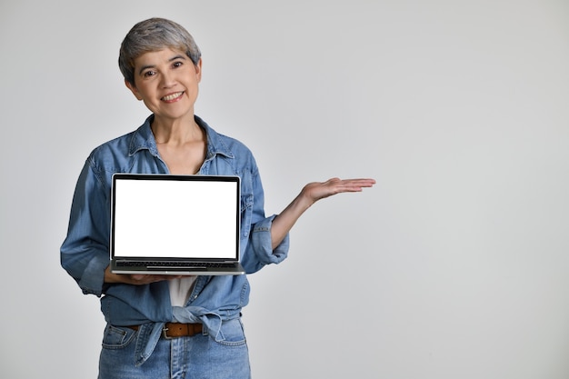 Ritratto di donna asiatica di mezza età anni '50 che indossa camicia di jeans casual t-shirt bianca che tiene il computer portatile con schermo spazio copia vuoto e palmo della mano aperta isolato su sfondo bianco