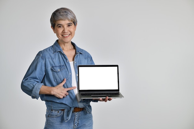 Ritratto di donna asiatica di mezza età anni '50 che indossa una camicia di jeans casual t-shirt bianca che tiene un computer portatile con schermo vuoto dello spazio della copia isolato su priorità bassa bianca. guardando la telecamera