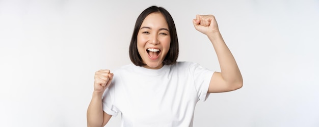 Ritratto di donna asiatica entusiasta che vince celebrando e trionfando alzando le mani per raggiungere l'obiettivo o il successo in piedi su sfondo bianco