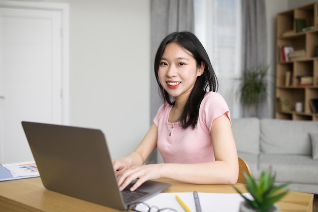 Ritratto di donna asiatica felice che utilizza il computer portatile seduto alla scrivania a casa e sorride allo spazio della copia della fotocamera