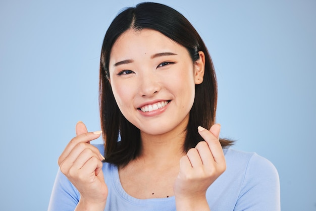 Ritratto di donna asiatica felice e segno d'amore o gesto della mano in romanticismo su uno sfondo blu studio Volto di persona femminile sorriso felice con amorevole icona emoji o simbolo per San Valentino