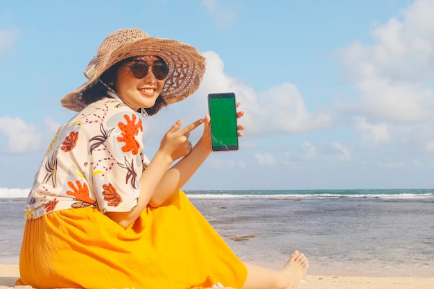 Ritratto di donna asiatica sorridente felice sulla spiaggia che mostra lo schermo verde di puntamento dello smartphone