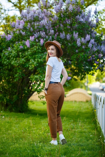 Ritratto di donna attraente alla moda in un cappello con lunghi capelli biondi nel parco, vestito con una maglietta bianca e pantaloni marroni.