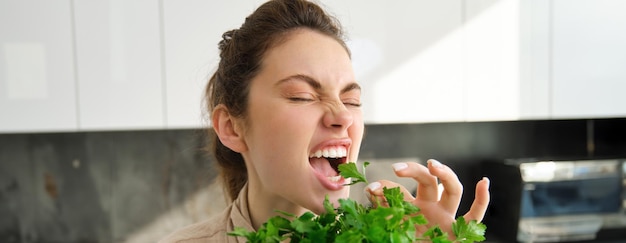 Ritratto di donna attraente che morde prezzemolo mangiando verdure fresche ed erbe in cucina