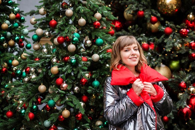 Ritratto di donna attraente in argento e cappotto rosso tra alberi di Natale con decorazioni festive.