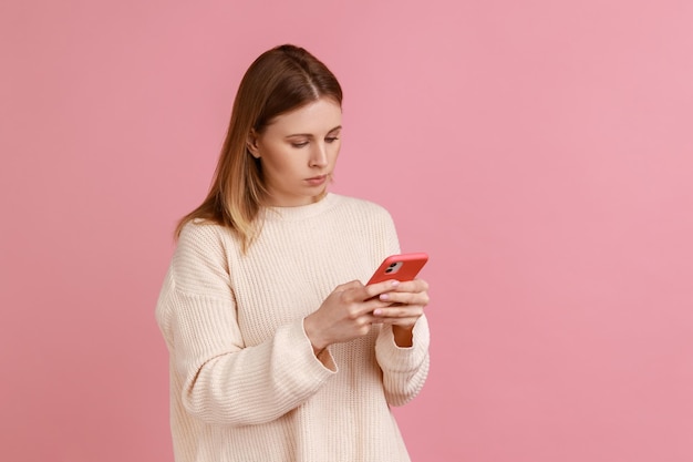 Ritratto di donna bionda attraente concentrata seria che utilizza il telefono cellulare guardando il messaggio di digitazione dello schermo del dispositivo che indossa un maglione bianco Studio indoor girato isolato su sfondo rosa