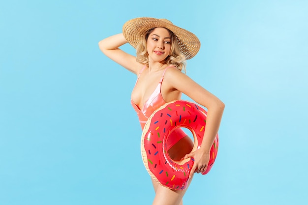 Ritratto di donna bionda attraente in costume da bagno e cappello da sole che tiene anello di gomma, in posa sexy alla telecamera. Stile di moda da spiaggia estiva, riposo in località di villeggiatura. girato in studio isolato su sfondo blu