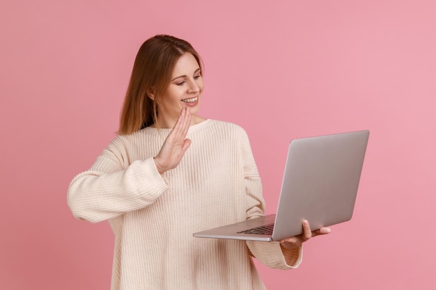 Ritratto di donna bionda attraente positiva che ha videochiamata agitando la mano alla telecamera dicendo ciao o arrivederci indossando un maglione bianco Studio indoor girato isolato su sfondo rosa