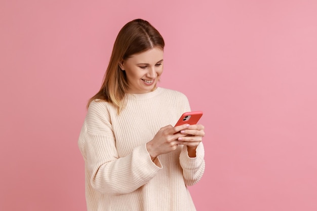 Ritratto di donna bionda attraente soddisfatta felice che utilizza smart phone, guardando lo schermo del dispositivo, scrivendo post, indossando un maglione bianco. Studio indoor girato isolato su sfondo rosa.