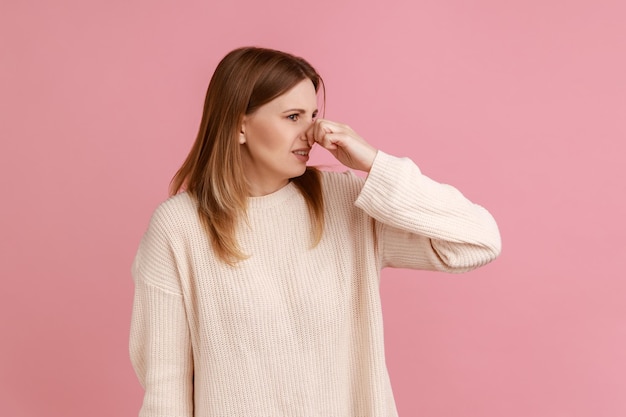 Ritratto di donna bionda con una smorfia di disgusto, trattenendo il respiro, pizzicando il naso con le dita per evitare cattivi odori, orribili odori, indossando un maglione bianco. Studio indoor girato isolato su sfondo rosa.