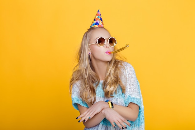 Ritratto di donna bionda divertente in cappello di compleanno e camicia blu su sfondo giallo celebrazione e festa