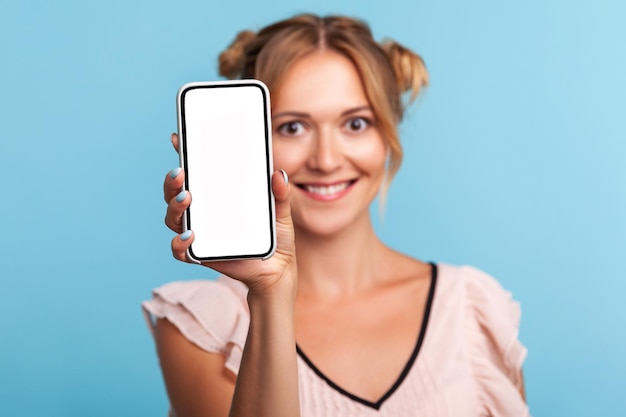 Ritratto di donna bionda in piedi con un sorriso affascinante, un'espressione felice e mostrando dispositivo mobile, telefono cellulare con display vuoto mock up per pubblicizzare. Studio indoor girato isolato su sfondo blu.