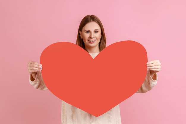 Ritratto di donna bionda romantica felice che tiene grande cuore rosso alla macchina fotografica ed esprime amore ed emozioni positive, indossando un maglione bianco. Studio indoor girato isolato su sfondo rosa.