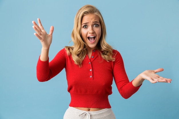 Ritratto di donna bionda sorpresa 20s indossando la camicia rossa alzando le mani, isolato sopra la parete blu