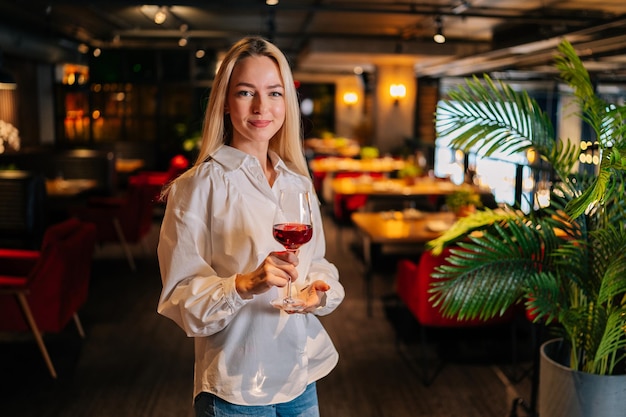 Ritratto di donna bionda sorridente che tiene un bicchiere di vino rosso in piedi nel ristorante con interni di lusso che guarda l'obbiettivo