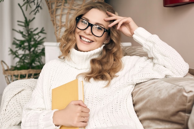 Ritratto di donna bionda splendida in maglione di lana bianco e occhiali da lettura libro in interni decorati luminosi accoglienti