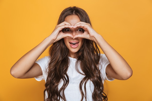 Ritratto di donna bruna 20s con i capelli lunghi sorridente e guardando attraverso la forma del cuore realizzato con le dita, isolato sopra il muro giallo