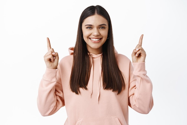 Ritratto di donna bruna allegra ed eccitata sorridente che punta il dito verso l'alto sul logo in alto che mostra la pubblicità in piedi su sfondo bianco in felpa con cappuccio
